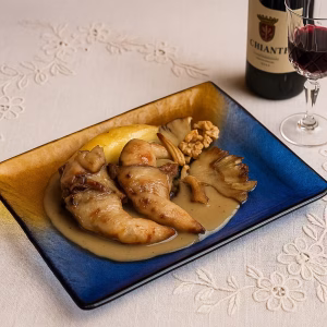 Perdiz estofada con peras y setas servida en plato rectangular de vidrio ámbar y azul sobre mantel bordado, junto a botella de vino Chianti y copa de cristal de Bohemia.
